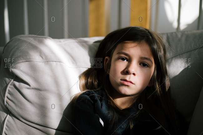 Little Girl With Dark Brown Hair Sitting On Sofa In Sunlight