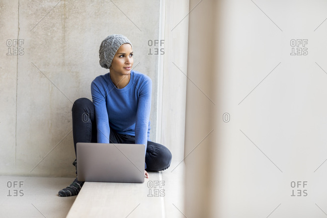 Portrait of young woman with laptop sitting on window sill looking out of window