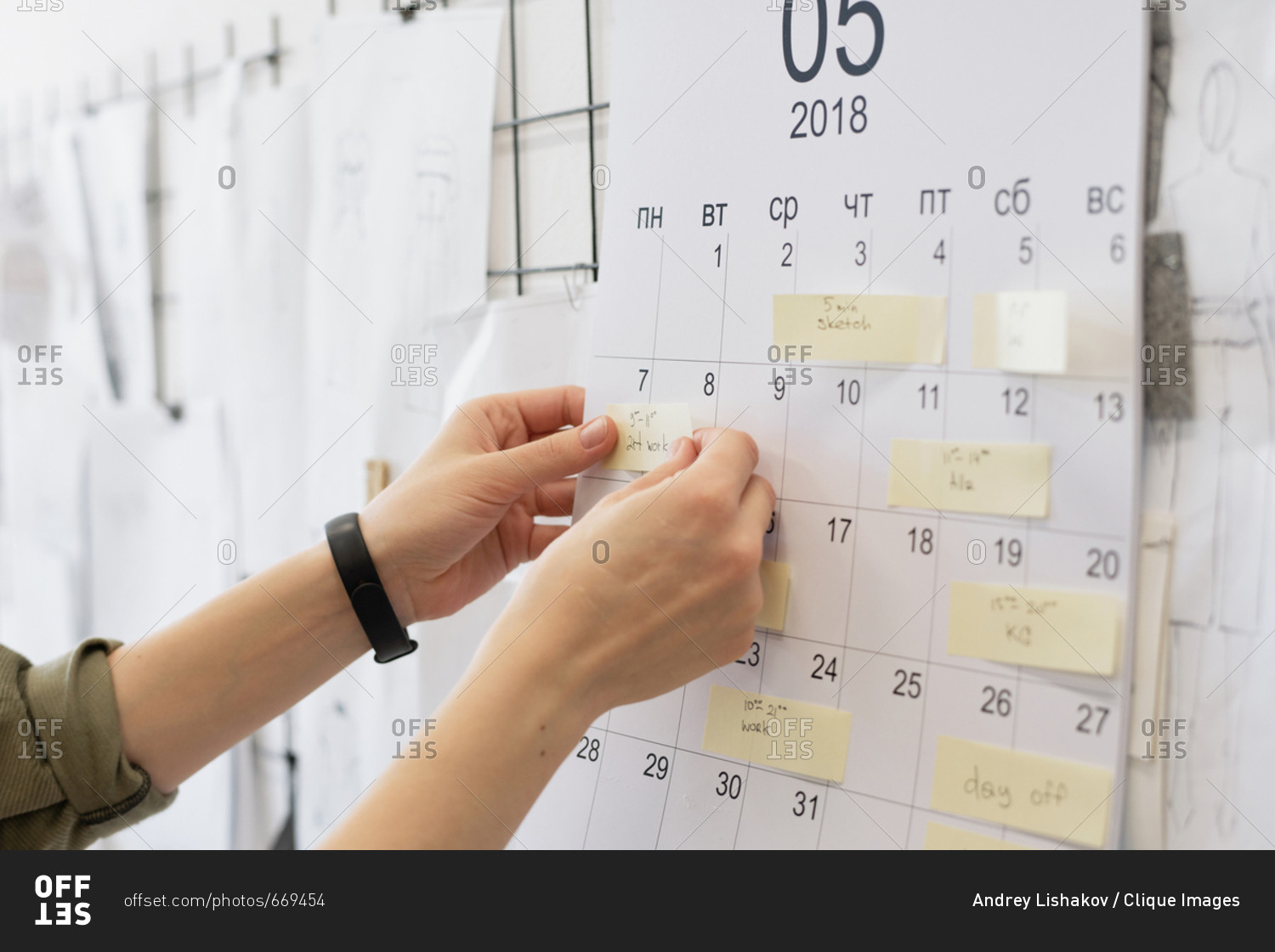 Close-up view of unrecognizable female fashion designer looking at sticky notes with plans on calendar hanging on wall