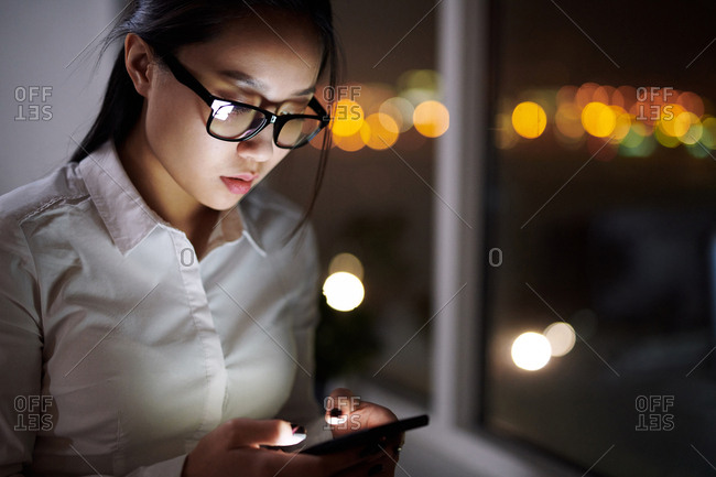 Asian corporate worker texting on Smartphone while standing by office window at night