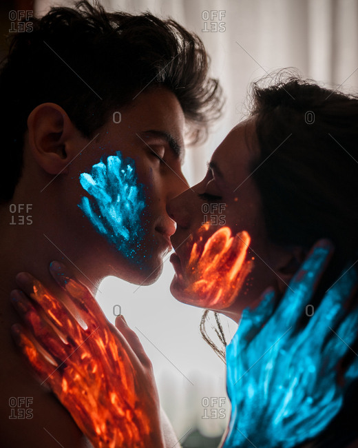 Young couple with orange and blue luminous paint kissing with eyes closed.