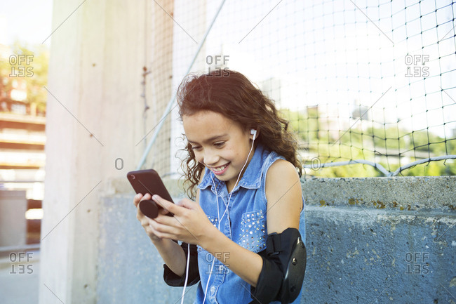Smiling girl with elbow pads and headphones listening music through smart phone in city