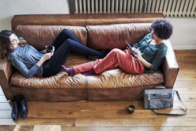 Two Young Women Lying On Couch With Cup Of Coffee And Tablet Stock Photo Offset