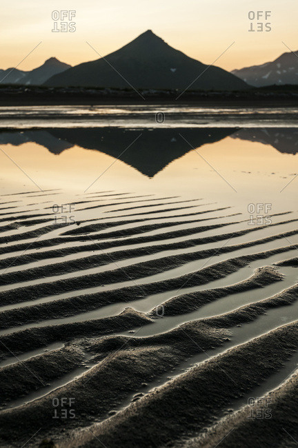 Alaska, United States of America - July 11, 2009: Silhouette Of A Peaked Mountain Reflected In Tranquil Water And Ripples In The Sand At Sunset; Alaska, United States Of America