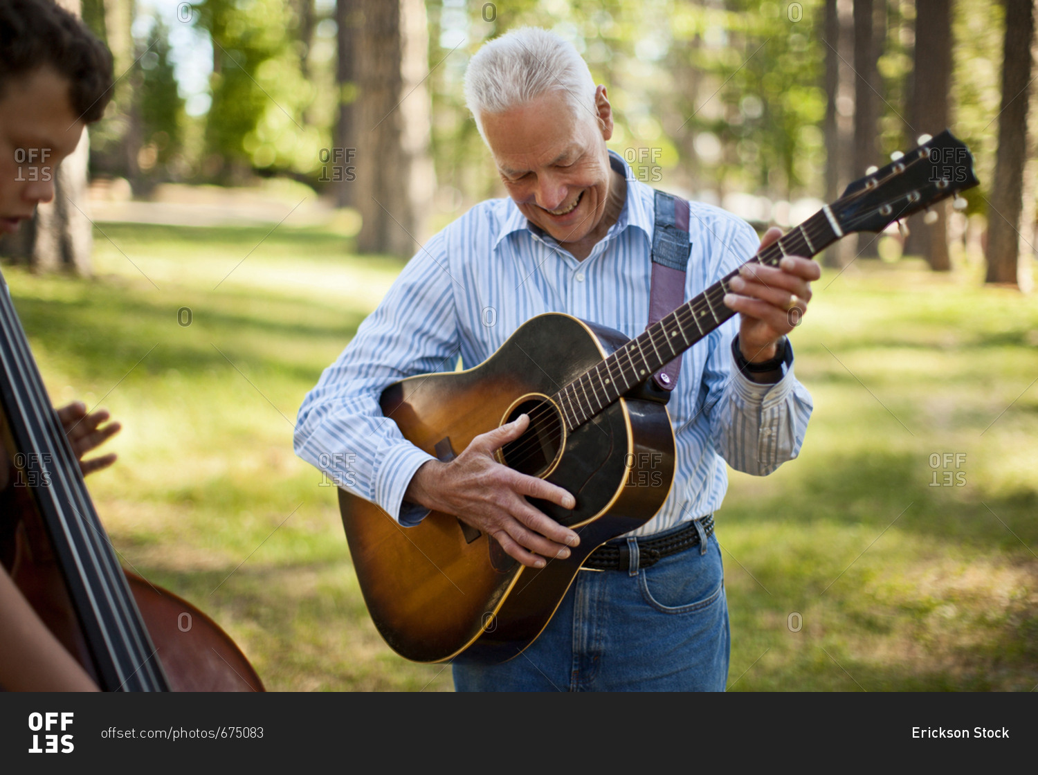 Smiling senior man playing an acoustic guitar while his grandson plays a double bass in a forest.