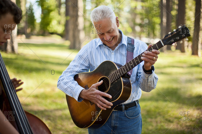 Smiling senior man playing an acoustic guitar while his grandson plays a double bass in a forest.