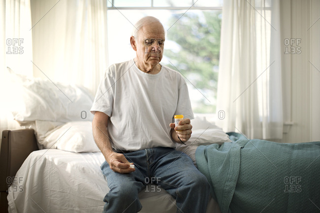 Concerned senior man holding a medicine bottle while sitting on the edge of his bed.