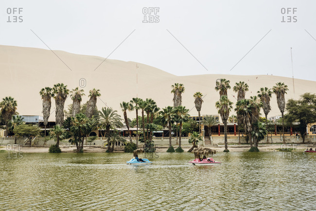 Huacachina, Peru - November 27, 2017: Paddleboats in green desert oasis lake in front of sand dunes