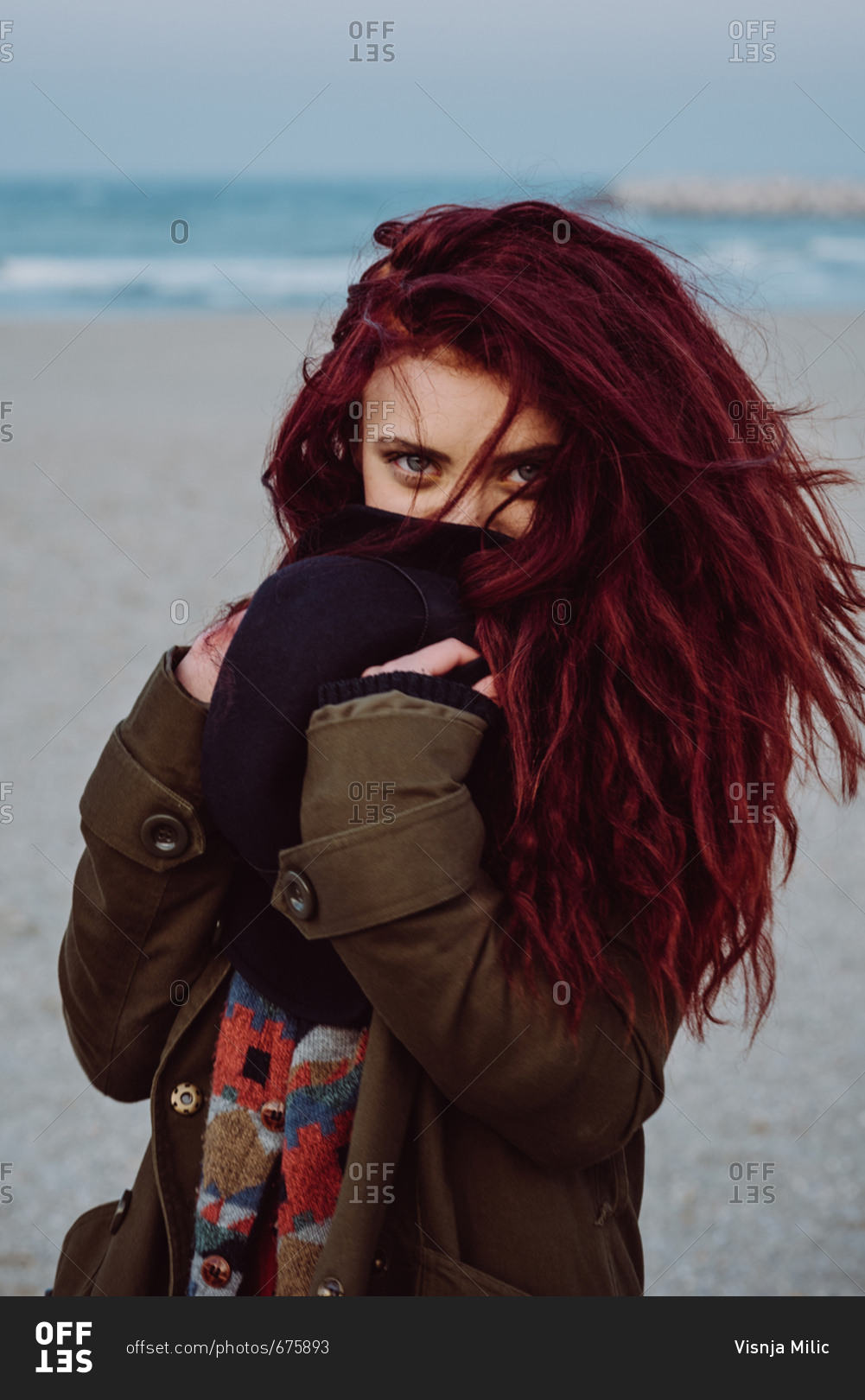 Teenager girl with long red hair walking on the beach wearing retro clothes covering her face with a hat