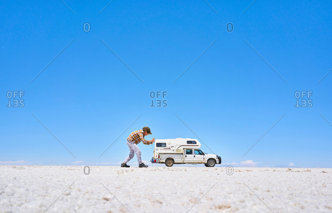 False perspective image of boy on salt flats, pretending to push recreational vehicle, vehicle in background, Salar de Uyuni, Uyuni, Oruro, Bolivia, South America