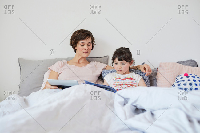 Mother and daughter relaxing in bed, daughter reading book, mother holding digital tablet