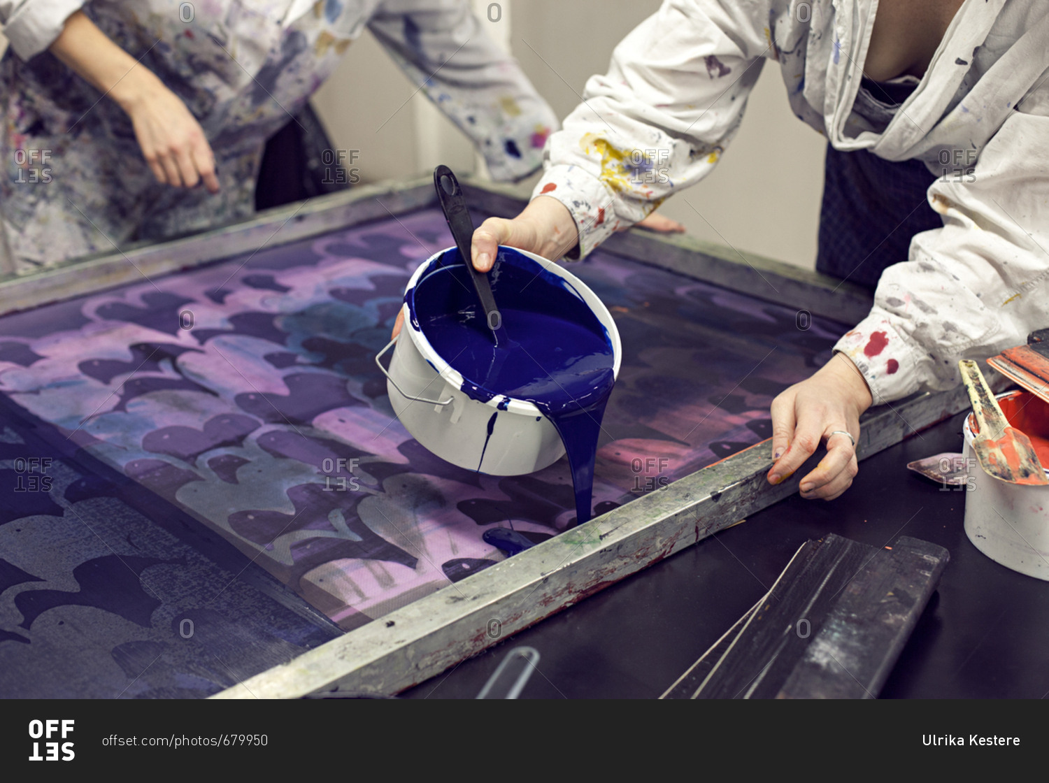Textile designer pouring ink onto stencil as partner holds screen print stencil in studio
