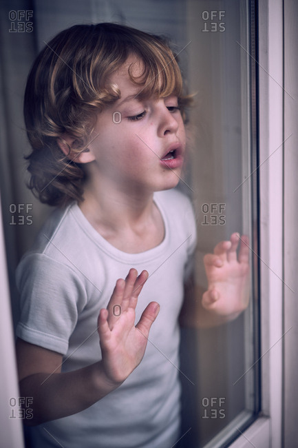 Cute little preschooler boy standing and breathing on window at home.