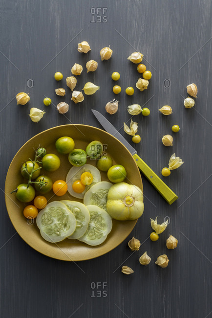 Husk cherries on a dark gray painted board with a bowl of cucumbers and cherry tomatoes