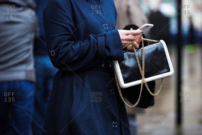 Woman using smart phone wearing navy trench coat holding black and white handbag with gold chain
