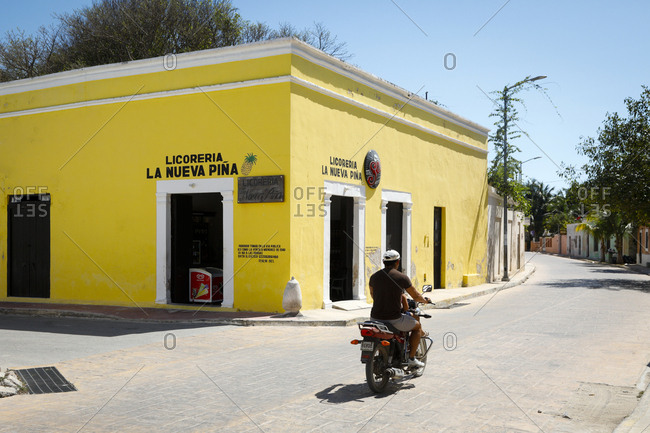 Celestun, Mexico - March 05, 2018: Man rides by colorful liquor store on motorbike
