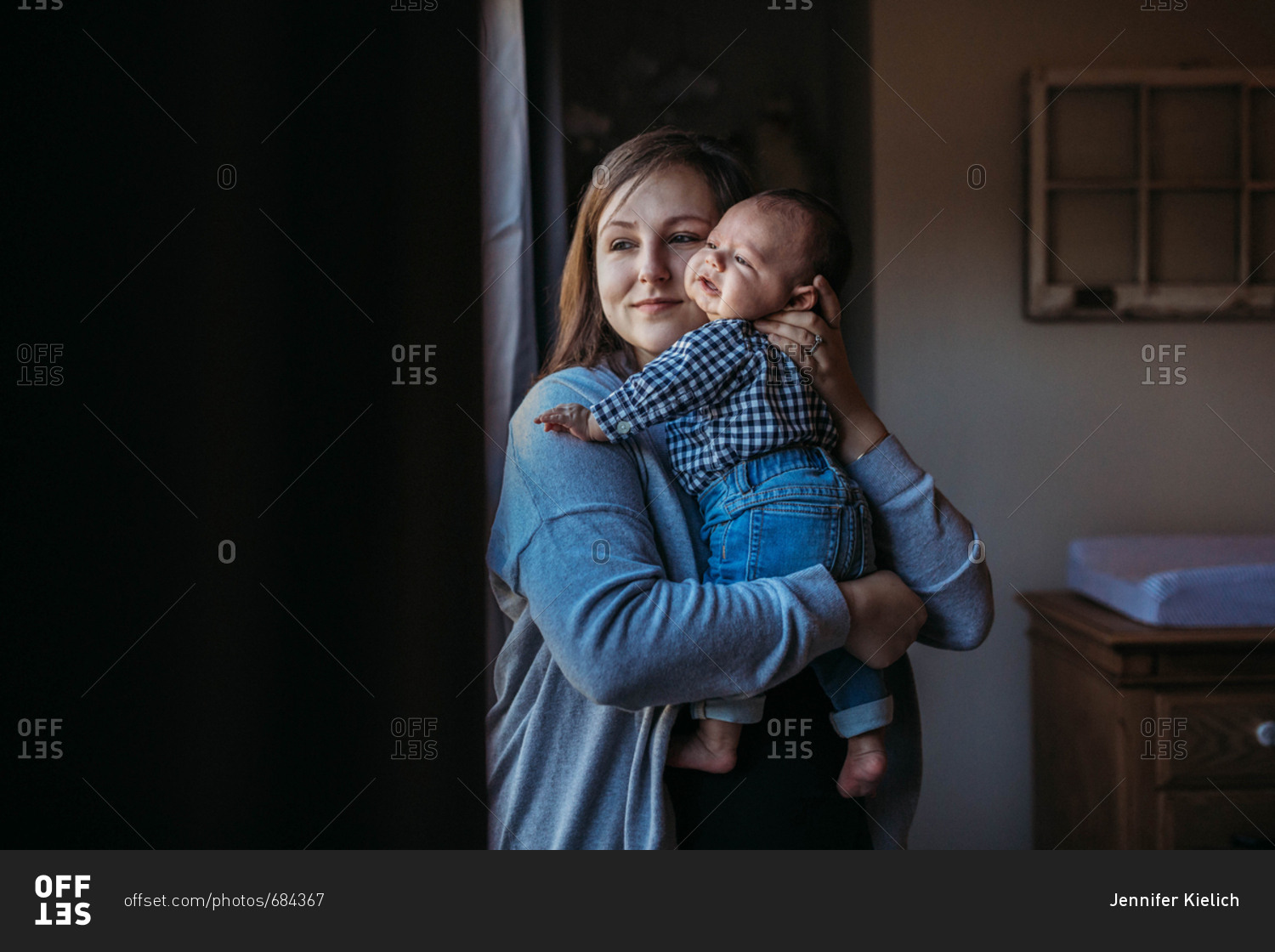 Mom holds newborn baby and stares out nursery window stock photo OFFSET
