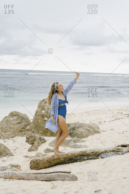 Young woman wearing vintage swim wear taking selfie with smartphone on beach in Bali, Indonesia