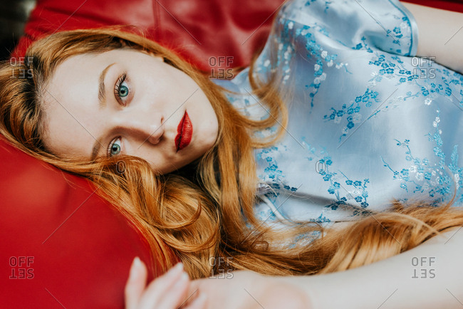Portrait of beautiful teenager girl with long red hair and blue eyes and pale skin laying on the red couch wearing blue dress and red lipstick
