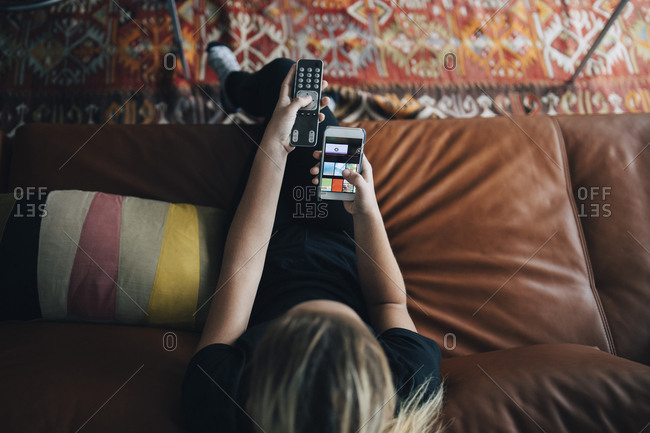 High angle view of teenage girl using phone app and remote control while sitting on sofa watching TV at home
