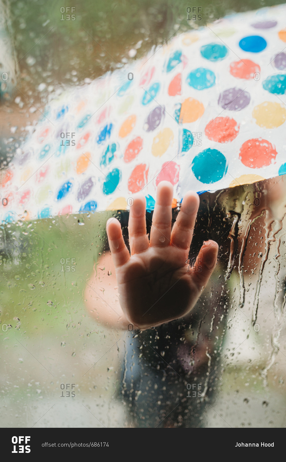 Little child holding umbrella standing outside in rain pressing hand against window