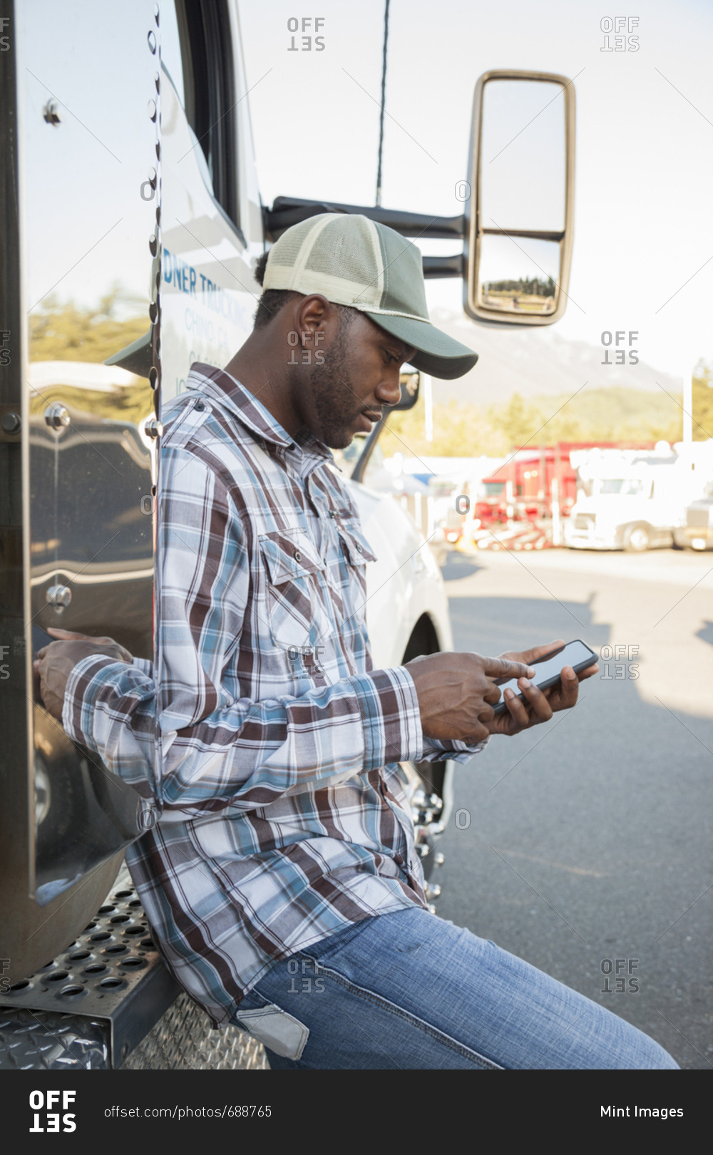Black man truck driver texting while standing next to his truck cab parked in a lot at a truck stop