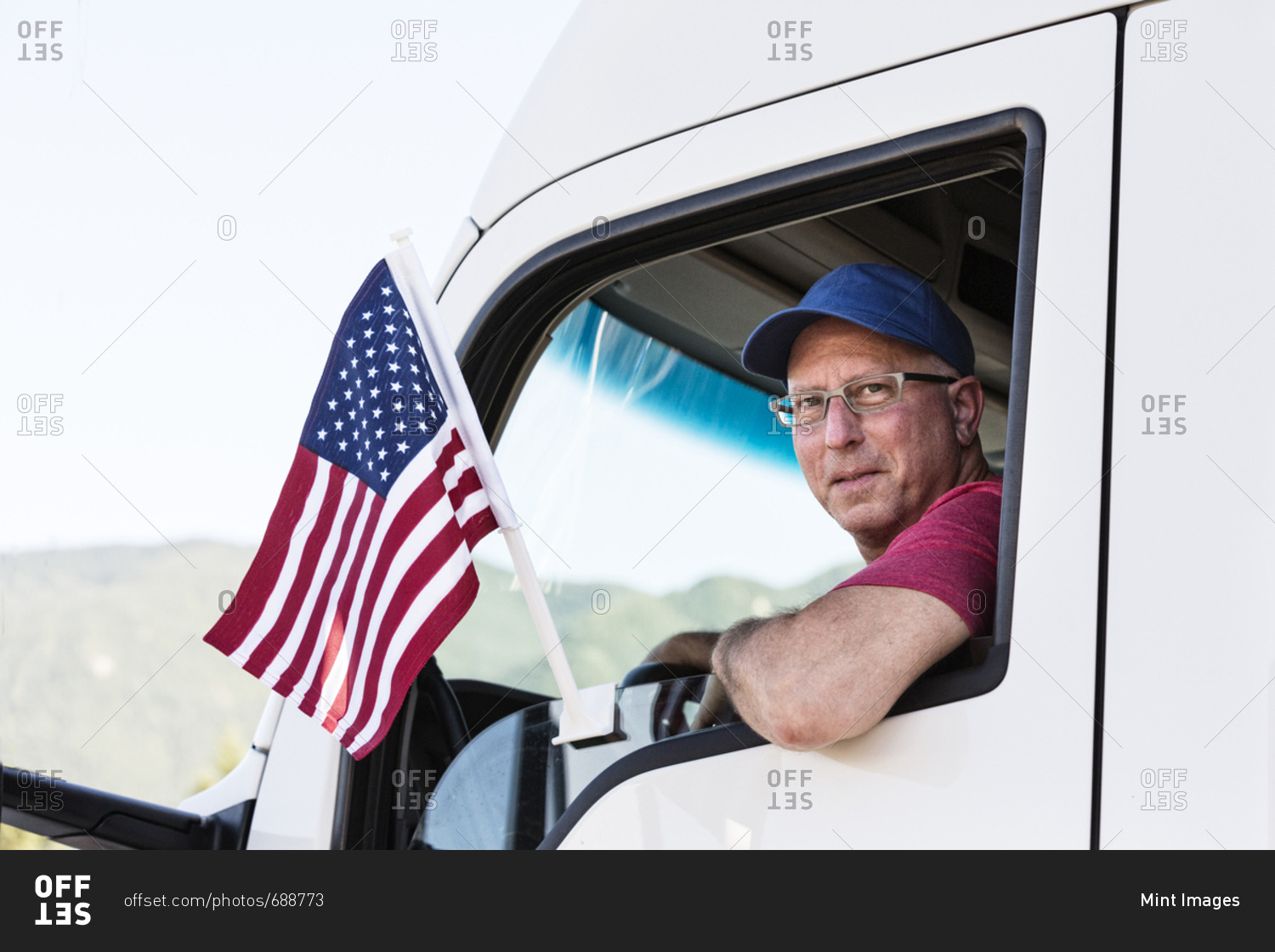 Caucasian man truck driver in the cab of his truck with an American Flag attached to the window