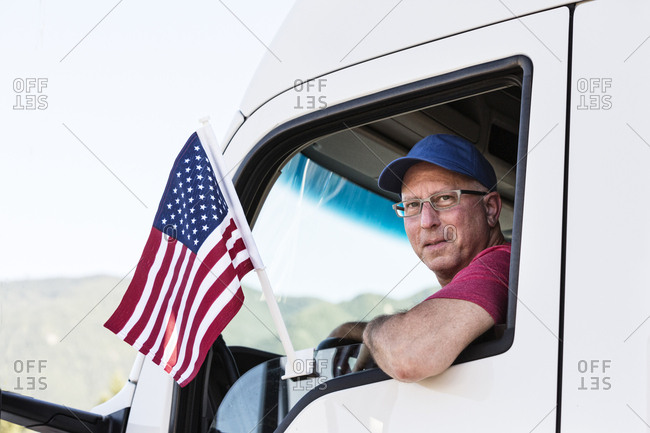 Caucasian man truck driver in the cab of his truck with an American Flag attached to the window
