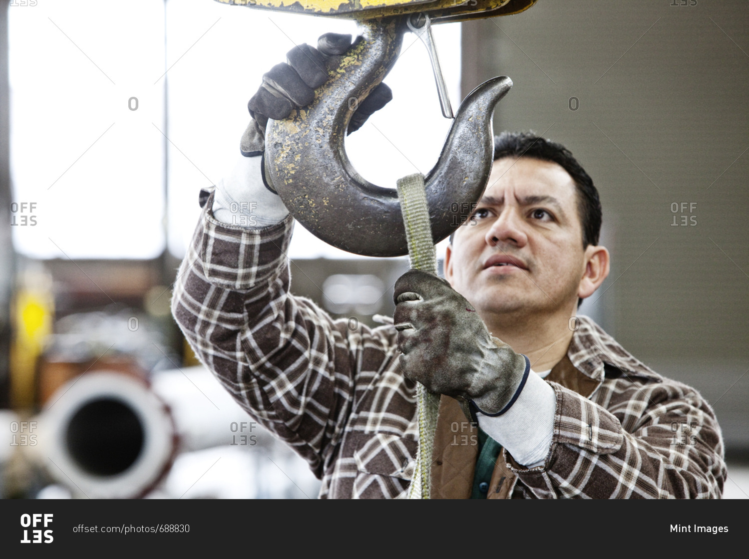 Hispanic man factory worker attaching a strap to the hook of a heavy duty overhead lift in a sheet metal factory