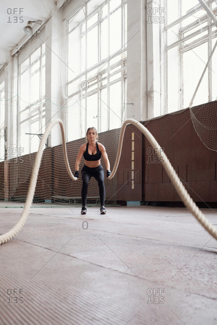 Young slim Caucasian woman in black sportswear burning calories by doing exercise with ropes in gym