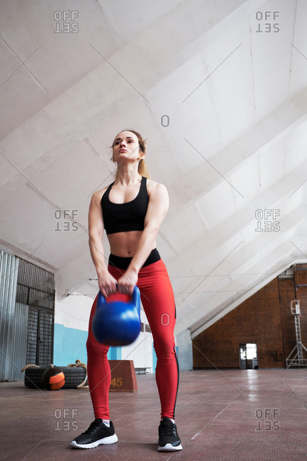 Low angle view of young Caucasian female athlete in sportswear doing exercises with kettlebell in old gym
