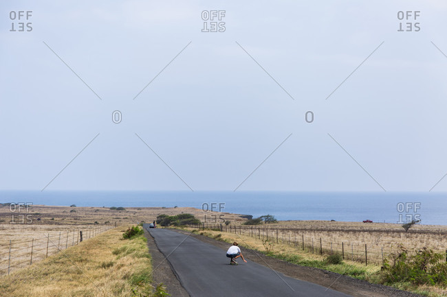 A man skateboards down the road towards Ka Lae, also known as South Point, the southernmost point of the Big Island of Hawaii and of the United States. People come to fish, jump off cliffs, and explore rock formations.