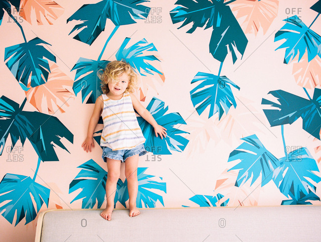 Little girl standing on bed against colorful wallpaper with palm leaf motif