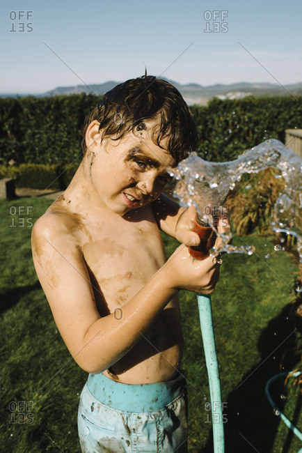 Dirty boy aiming water hose at camera on sunny day