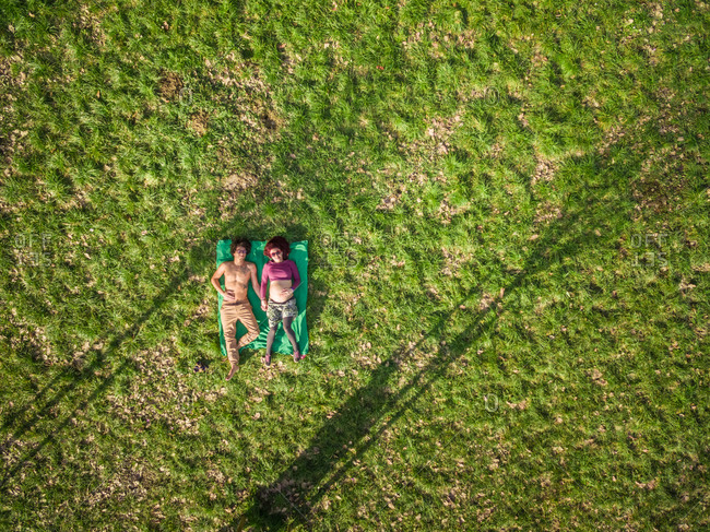 Aerial view of happy young man and pregnant woman holding hands while lying in grass at park