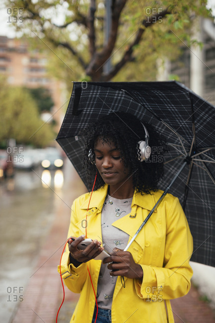 Young black woman listening to music on smartphone while waiting on side of street holding umbrella stock photo - OFFSET