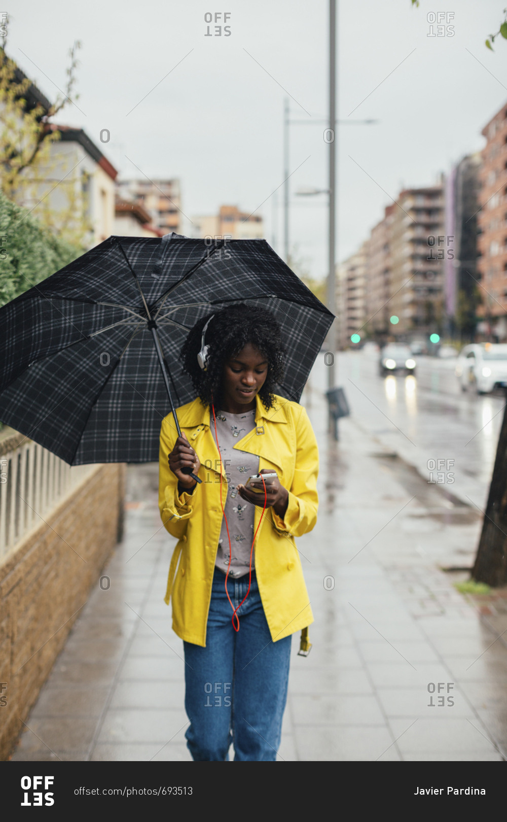 Young black woman looking down at phone while walking down the street in the rain