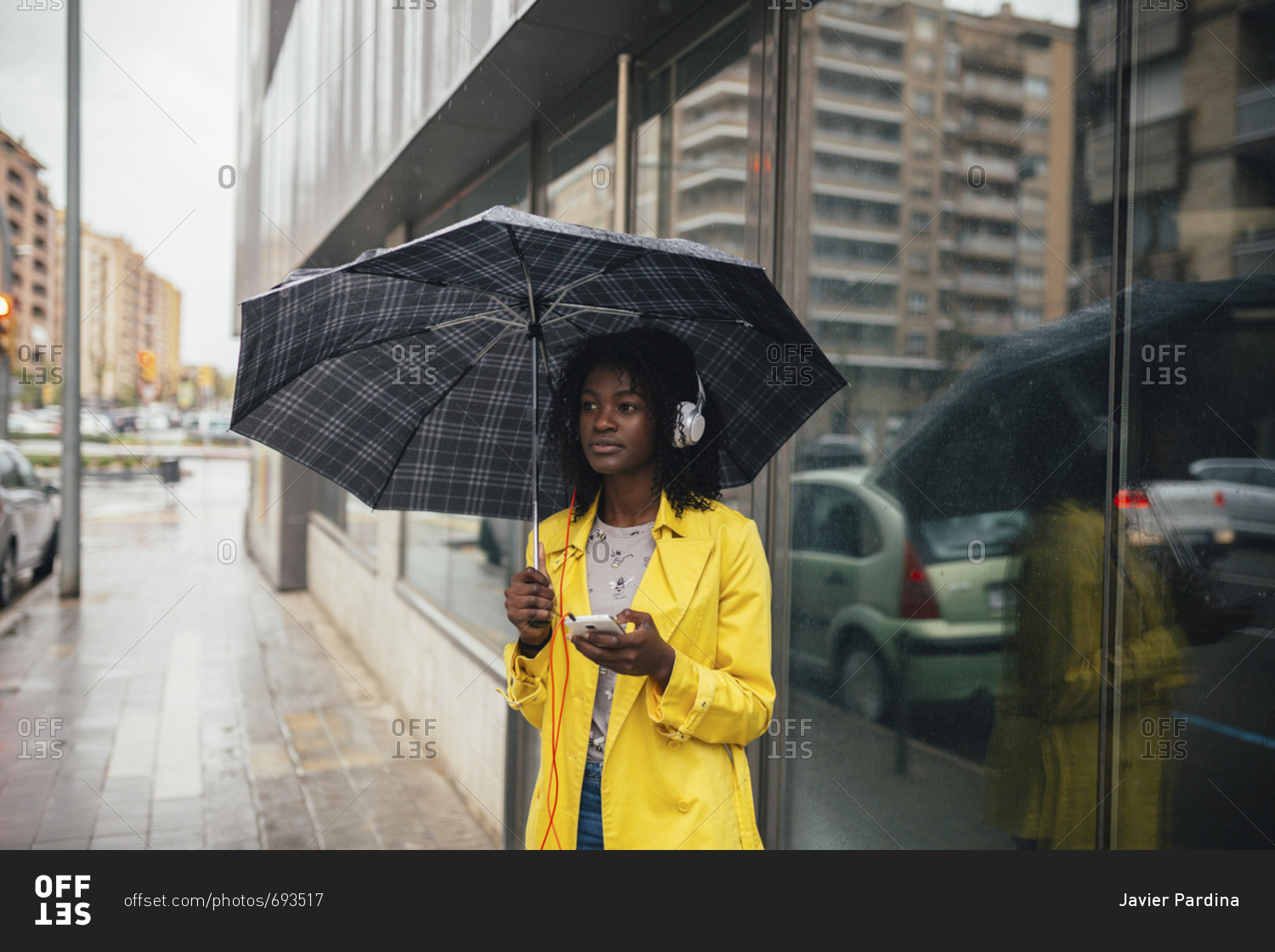 Young black woman waiting in the rain on the street while listening to music on smartphone