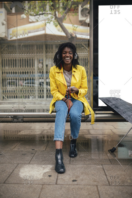 Young woman laughing while talking on smartphone in bus shelter on gloomy day