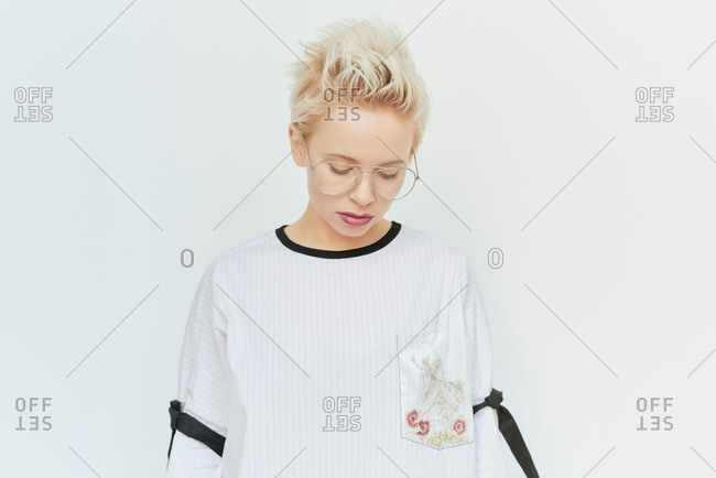 Portrait of young stylish woman with short blond hair wearing clear-lens sunglasses looking down on white background