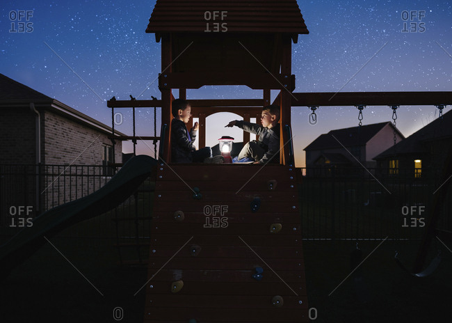 Brothers with illuminated lantern sitting on outdoor play equipment against star field at park
