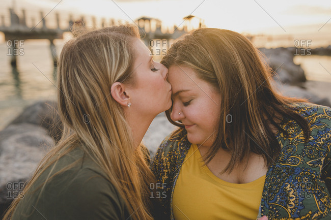 A lesbian woman kisses her lover's forehead at sunset near a pier