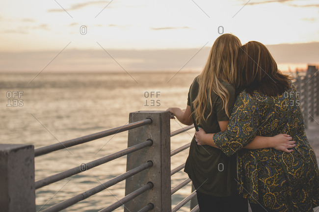 A lesbian couple stand arm in arm as they watch the sunset over the pier