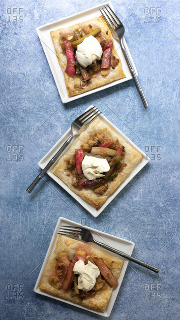 Three individual rhubarb and almond tarts with forks on a textured blue background.