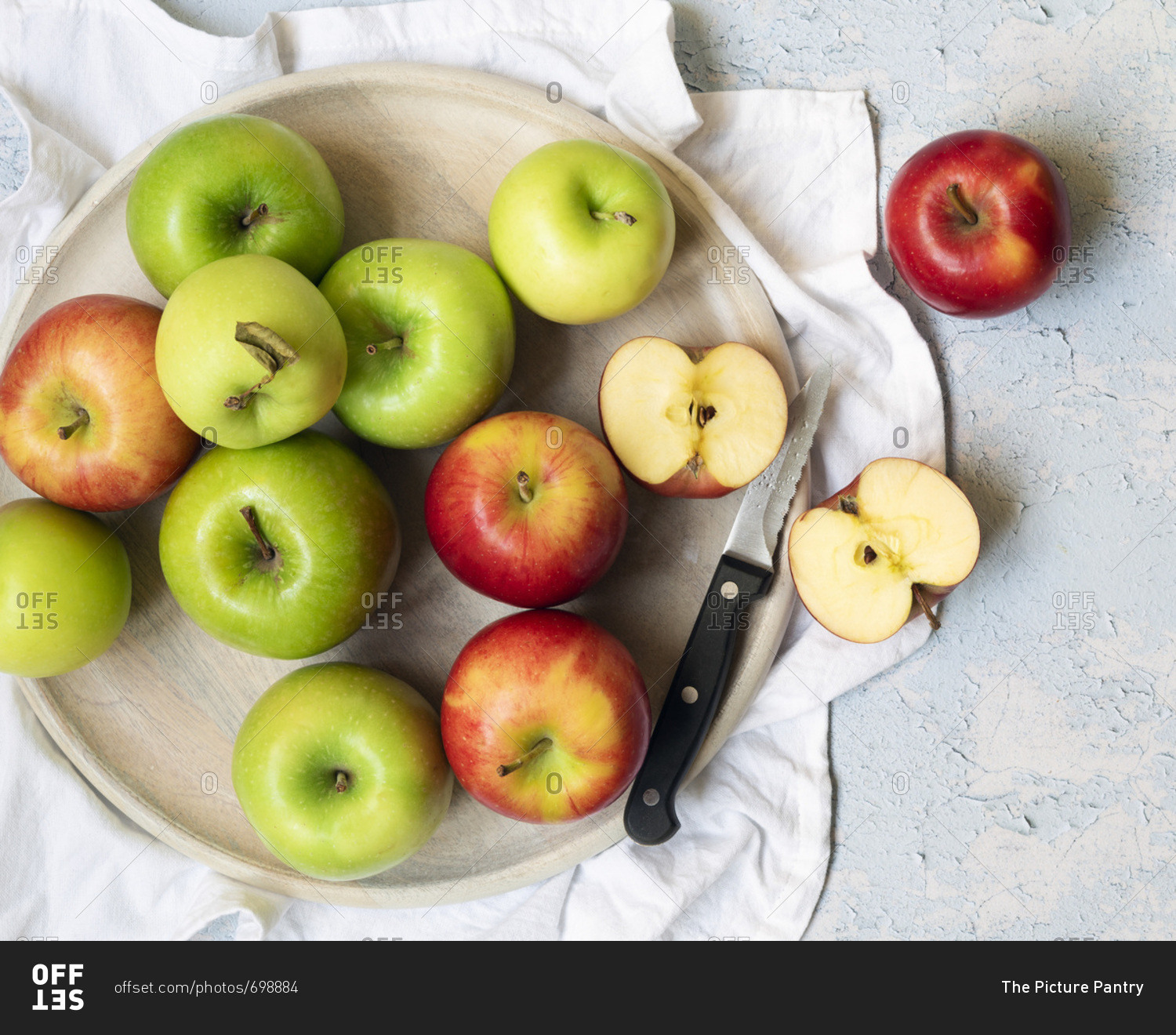 Green and red apples with two apple halves and knife on a round wooden serving board.
