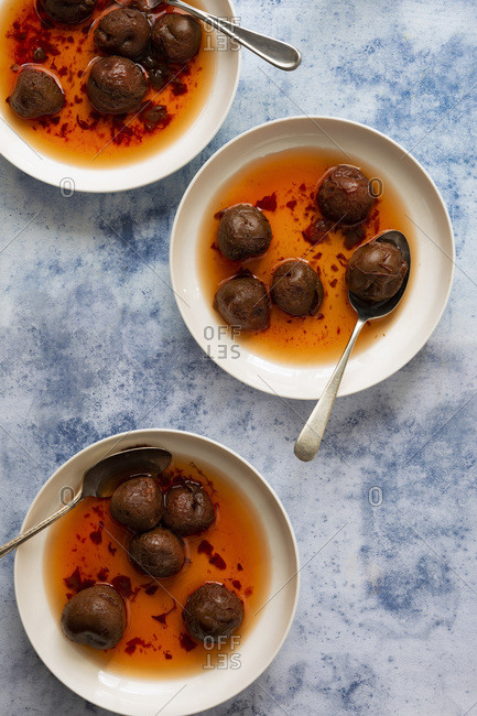 Three bowls of stewed red plums in syrup with spoons on a blue textured background.