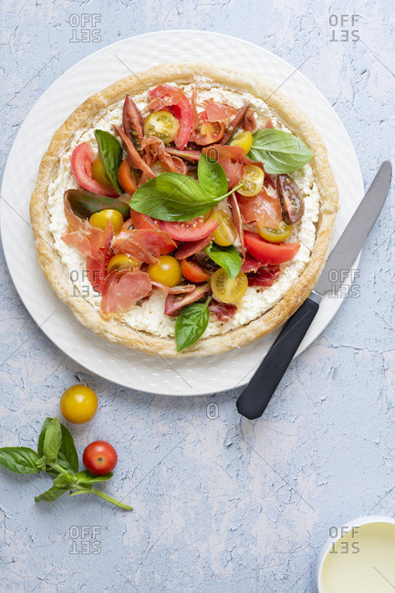 Round tart filled with cherry tomatoes, pancetta, feta cheese and basil on a plate with a knife.