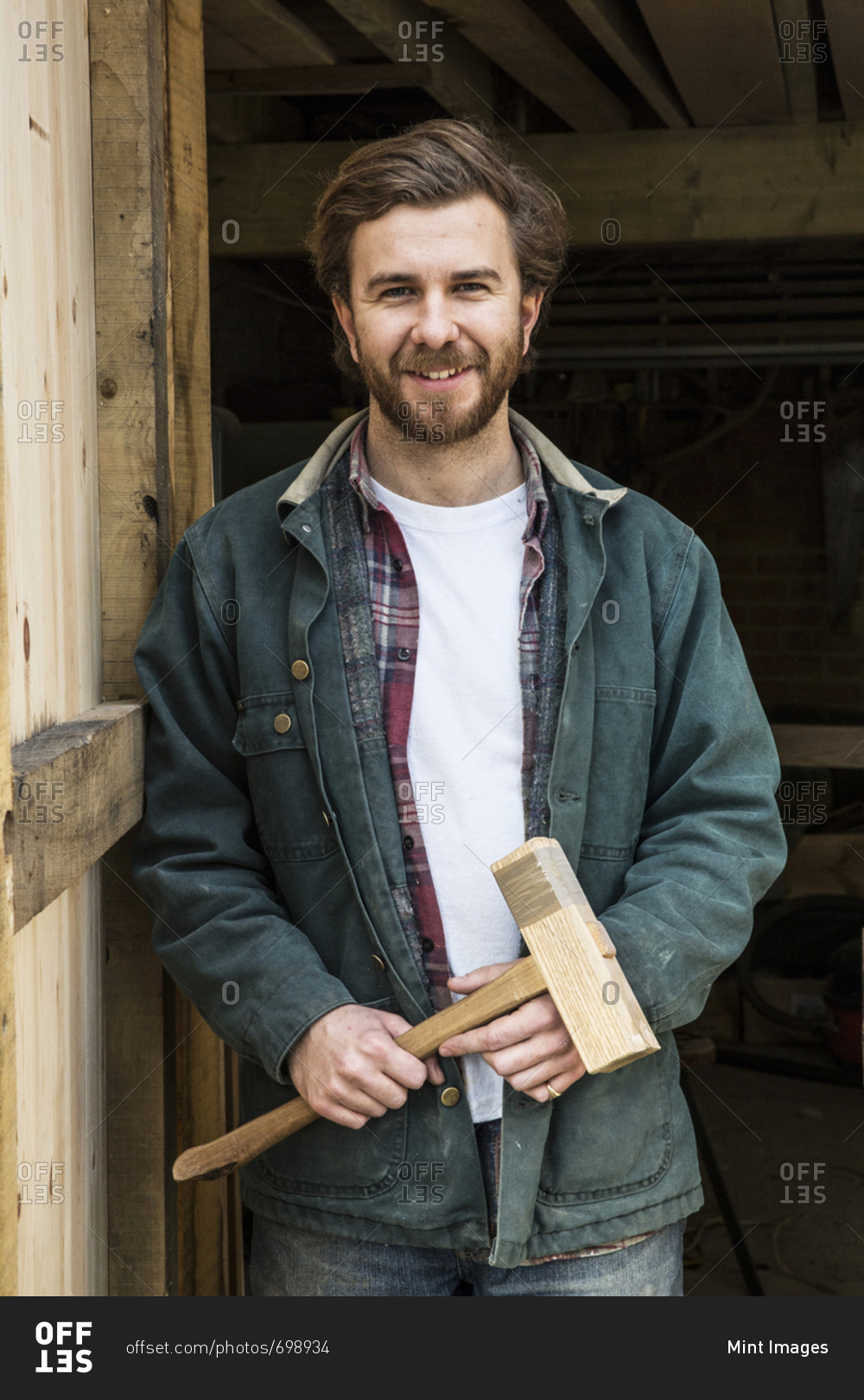 Bearded man standing in doorway of woodworking workshop, holding wooden mallet, smiling at camera