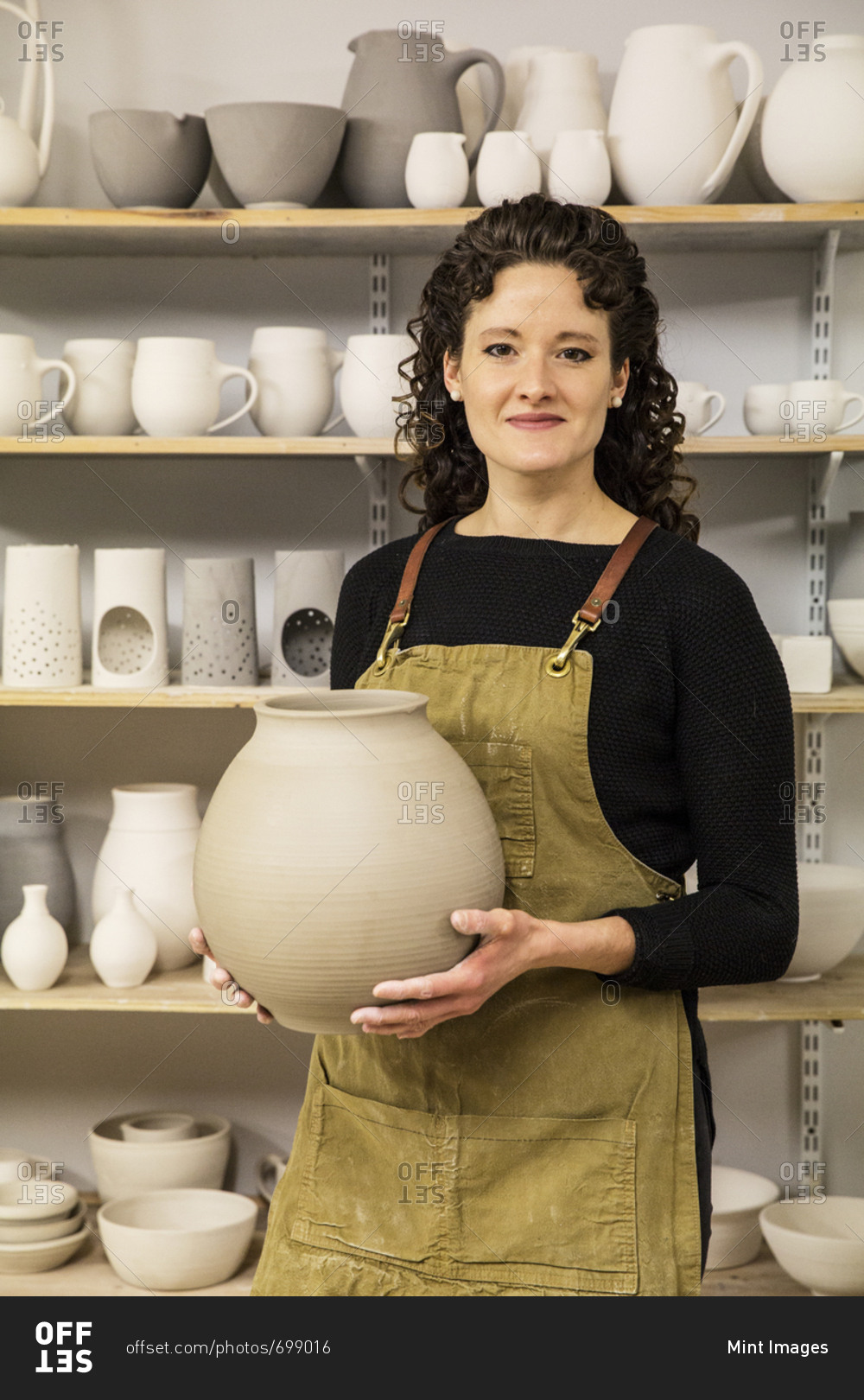 Woman with curly brown hair wearing apron holding unfired spherical clay vase