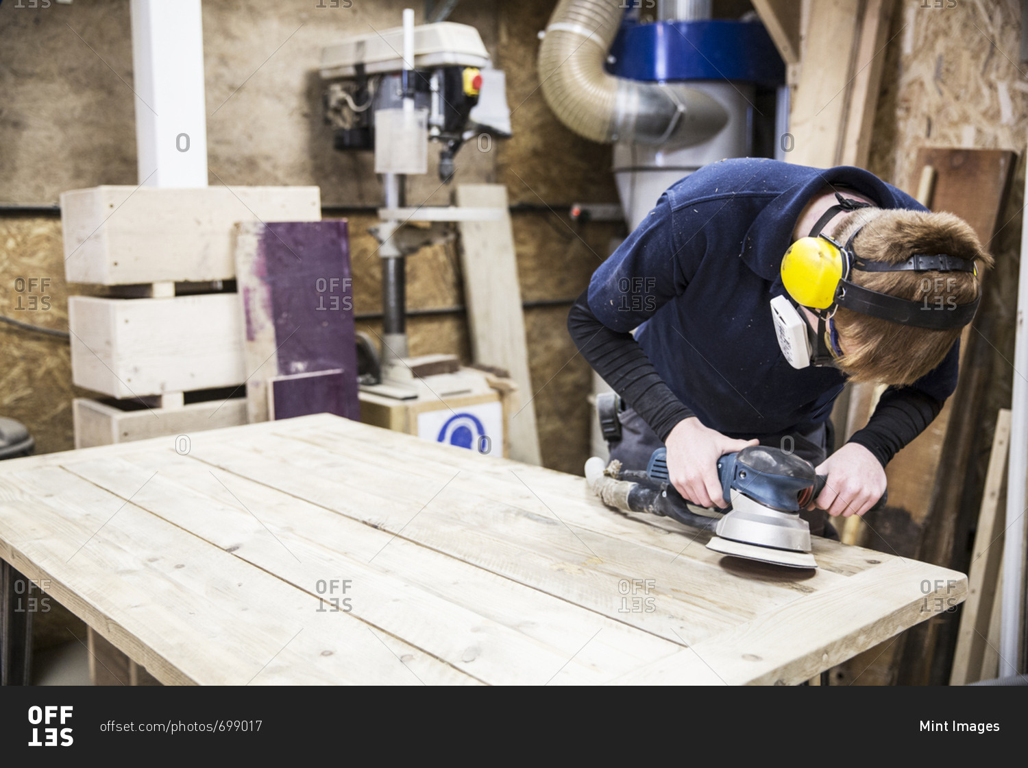 Man wearing ear protectors, protective goggles and dust mask standing in a warehouse, using sander to smoothen piece of wood